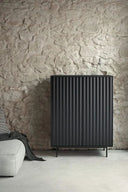 Mueble auxiliar alto SIERRA de 97 cm con 2 puertas y 2 cajones, en negro, en un salón con pared de piedra – Teulat